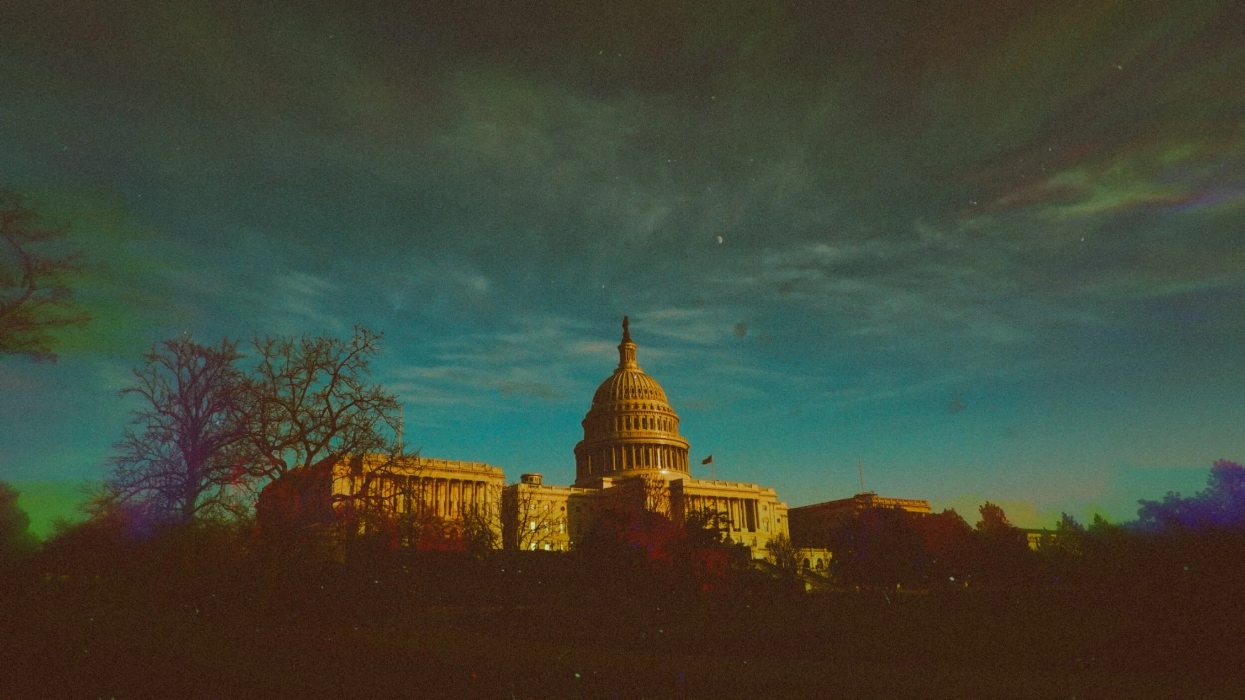 Night view of the U.S. Capitol building illuminated, with a starry sky and faint aurora in the background, and leafless trees in the foreground.