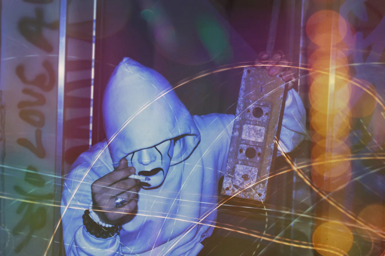 Person wearing a hoodie and a painted mask, holding a sparkler in one hand and a metal object in the other, with swirling light effects in the background.