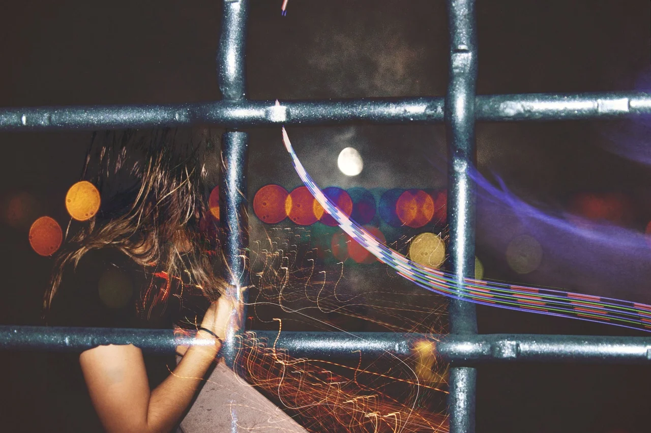 A girl with long hair holding a sparkler at night, seen through a metal fence with light trails and bokeh lights in the background