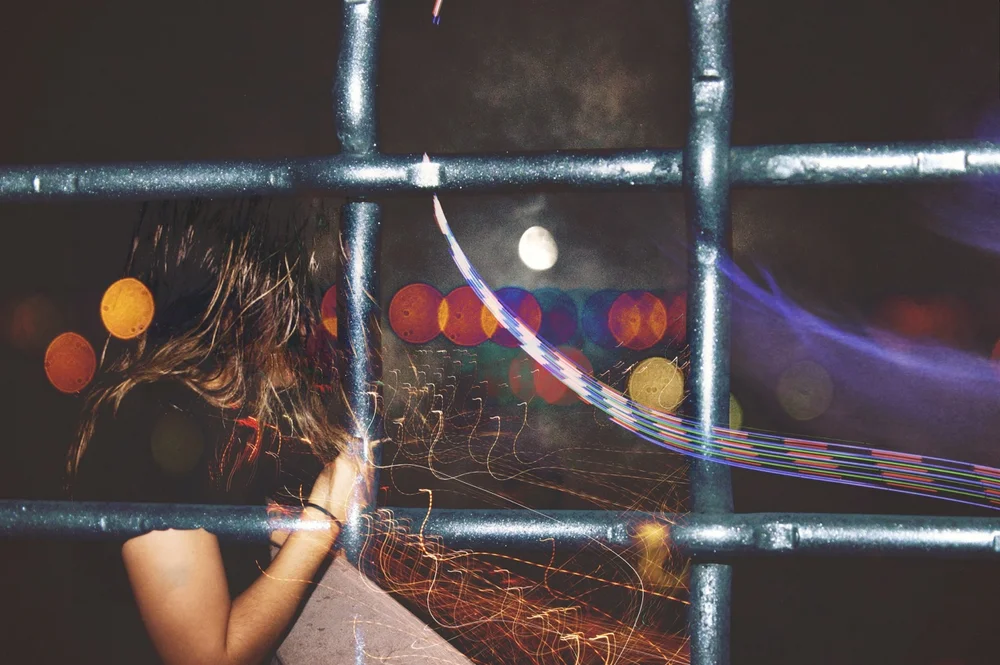 A girl seen through a metal fence at night, with bokeh lights behind her.