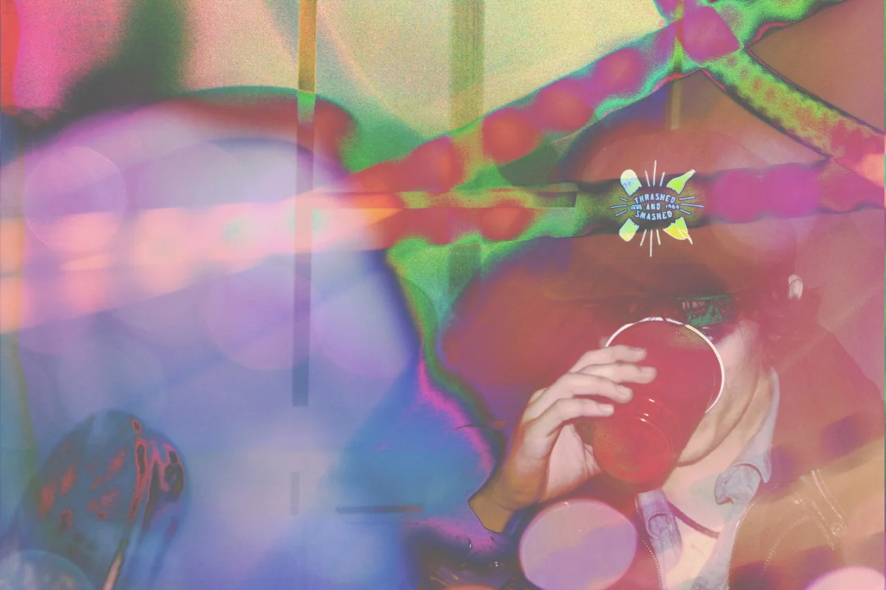 Person drinking from a red cup, wearing glasses with the word 'Transformers' on the glasses, and sitting in a colorful, abstract background with rainbow hues and bokeh effects.