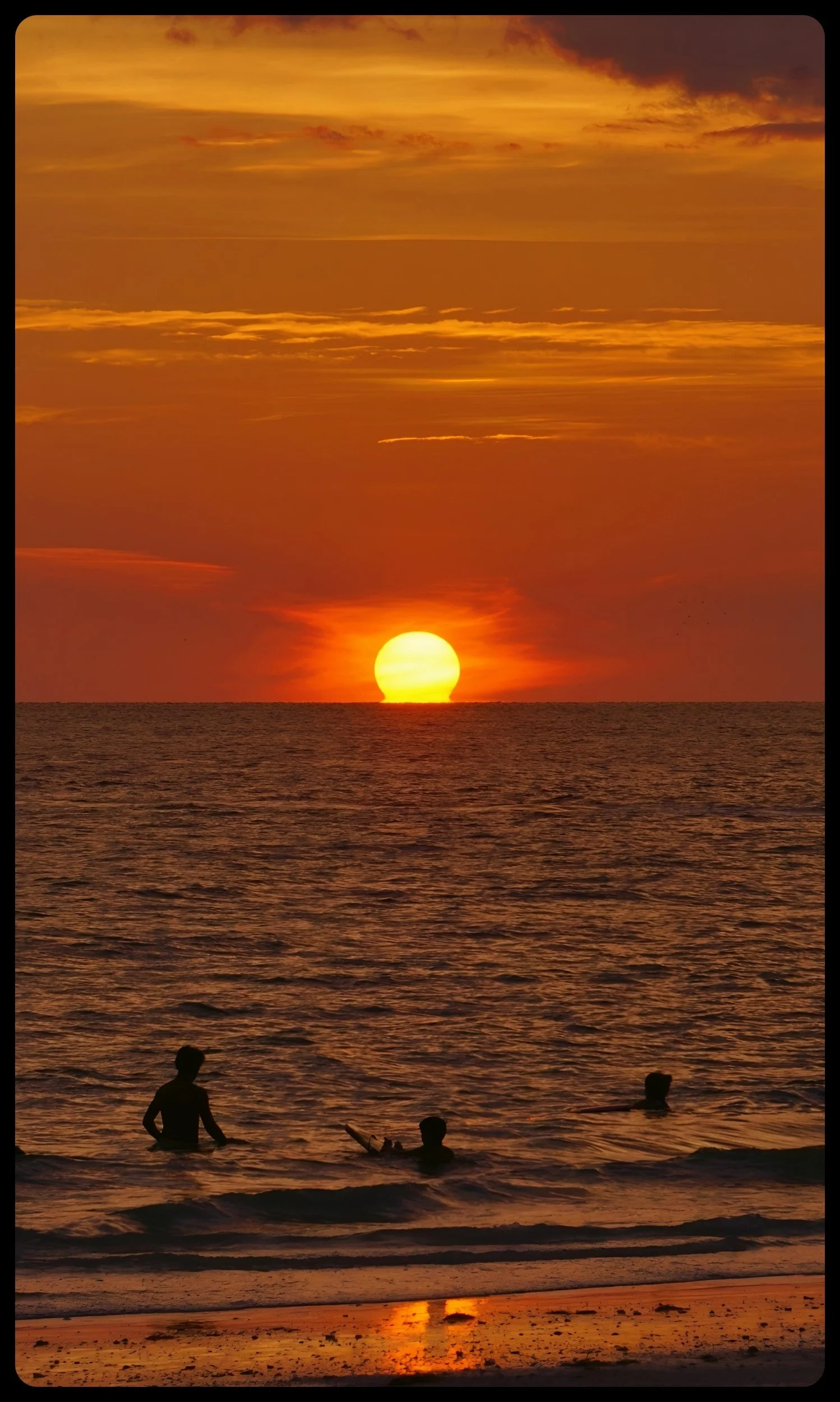 Sunset over the ocean with a few people swimming and playing in the water, silhouetted against the orange and yellow sky.