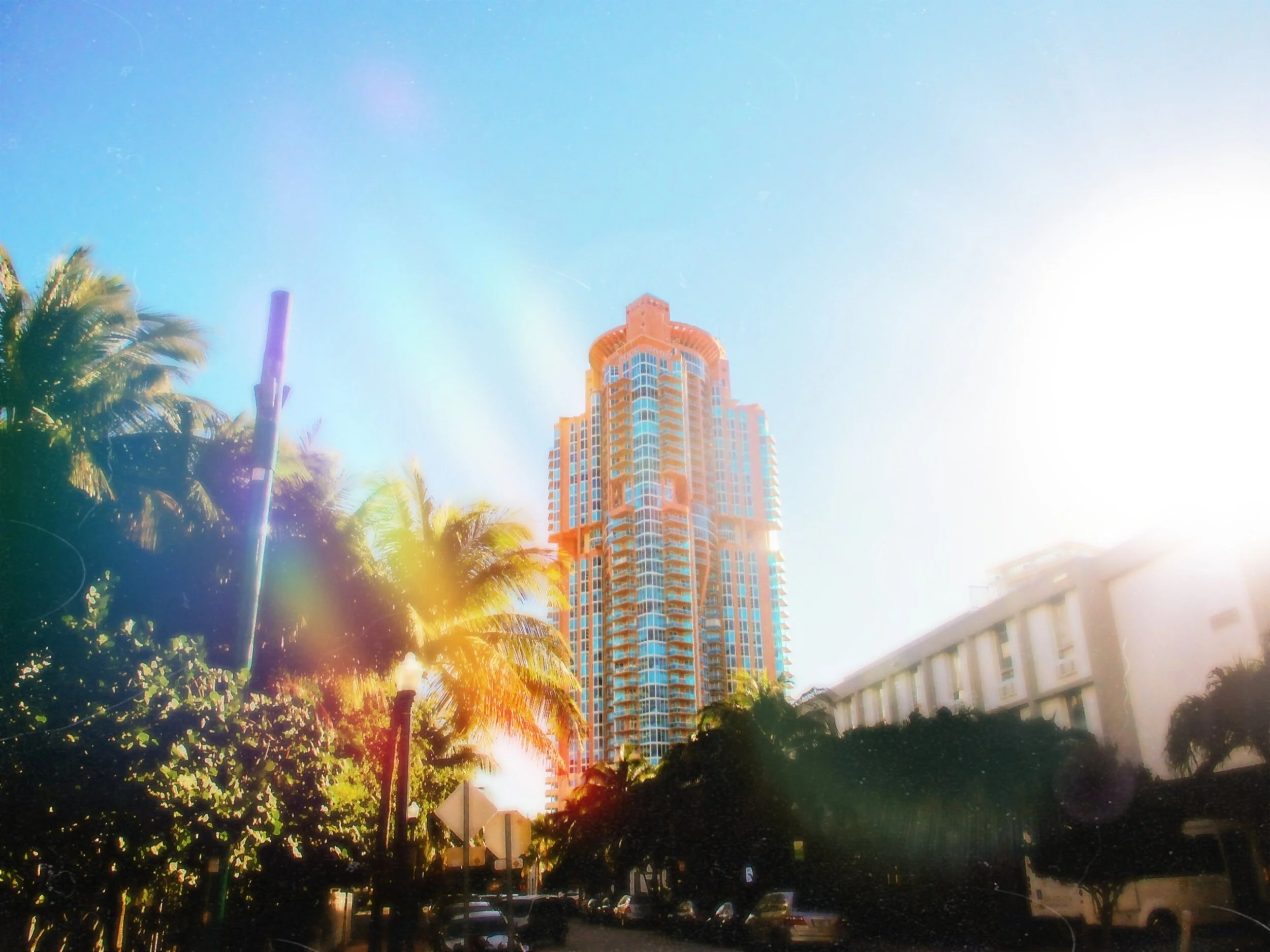 Tall high-rise building with balconies and a rounded top, surrounded by palm trees and parked cars, under a bright blue sky with sunlight.