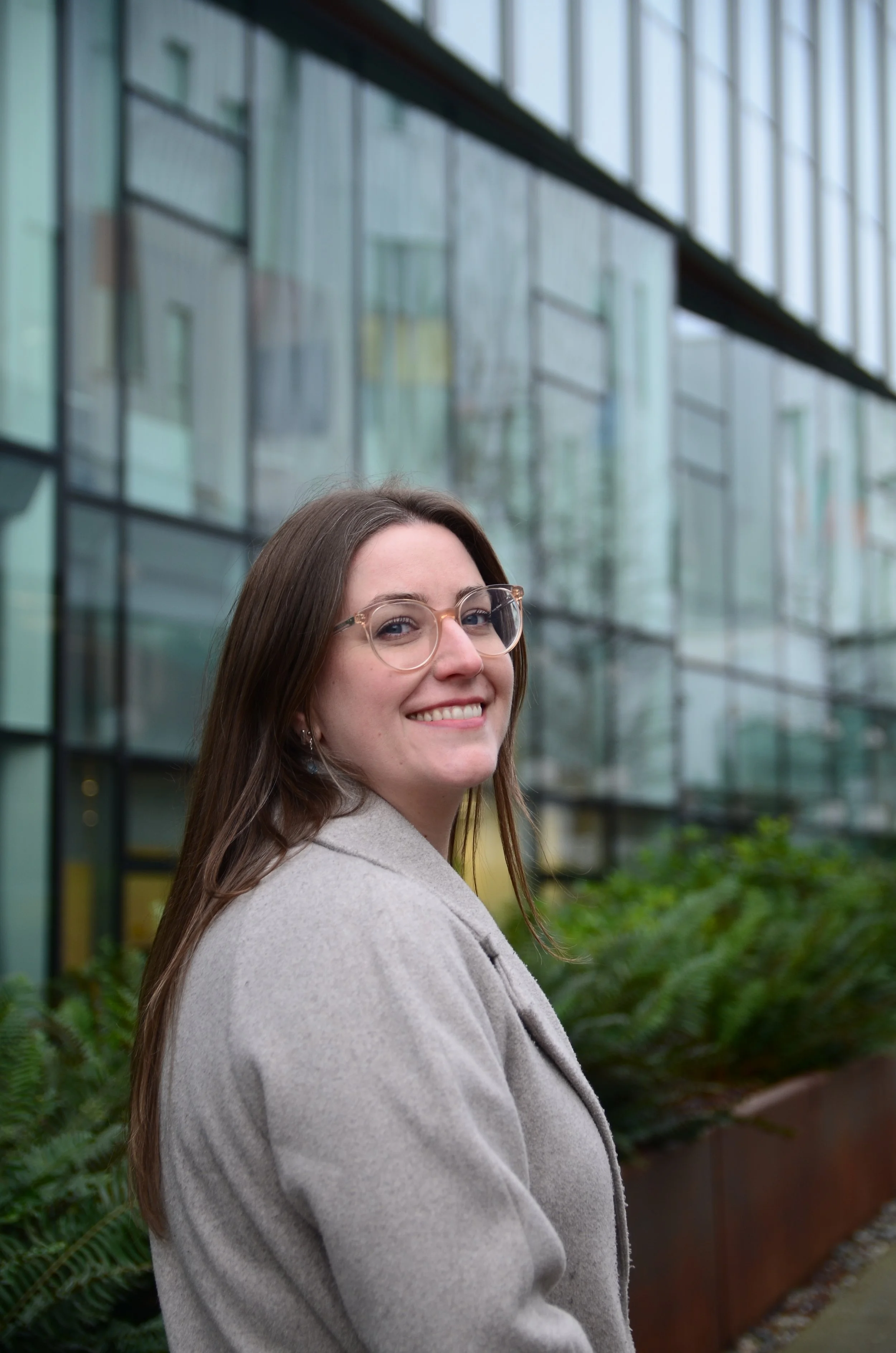 A woman with long brown hair, wearing glasses and a beige coat, smiling outdoors in front of a modern glass building.
