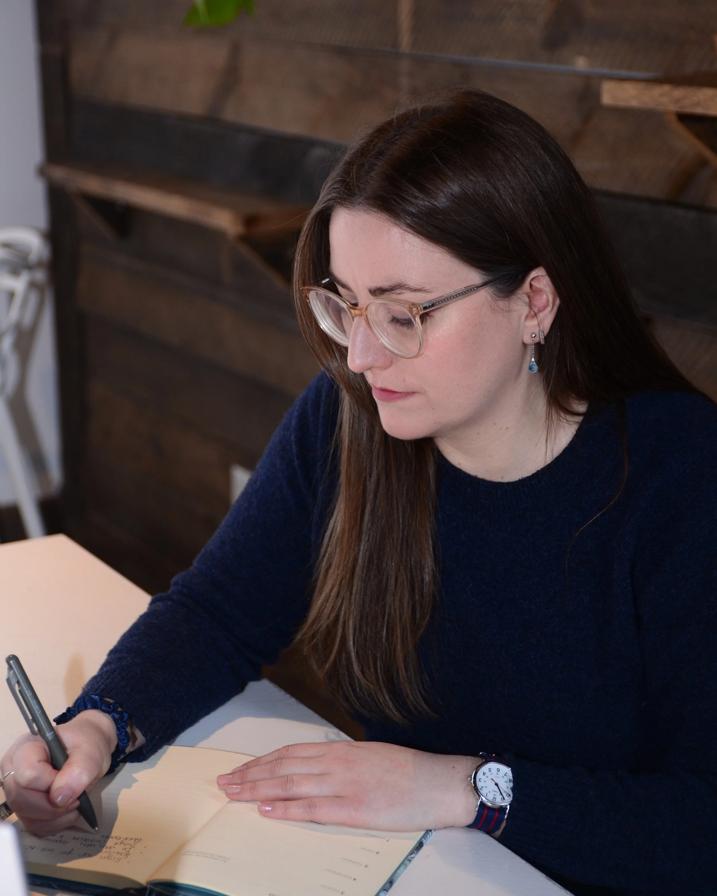 A woman with long brown hair, glasses, and earrings sitting at a desk writing in a notebook.