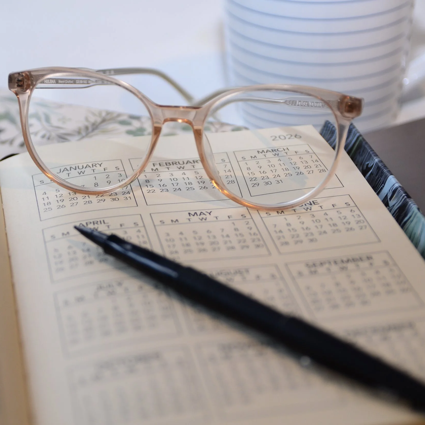 Blush coloured eyeglasses resting on a page of a 2026 calendar with a black pen nearby.