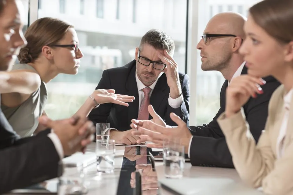 A group of professionals in a serious discussion around a table in a modern office conference room.