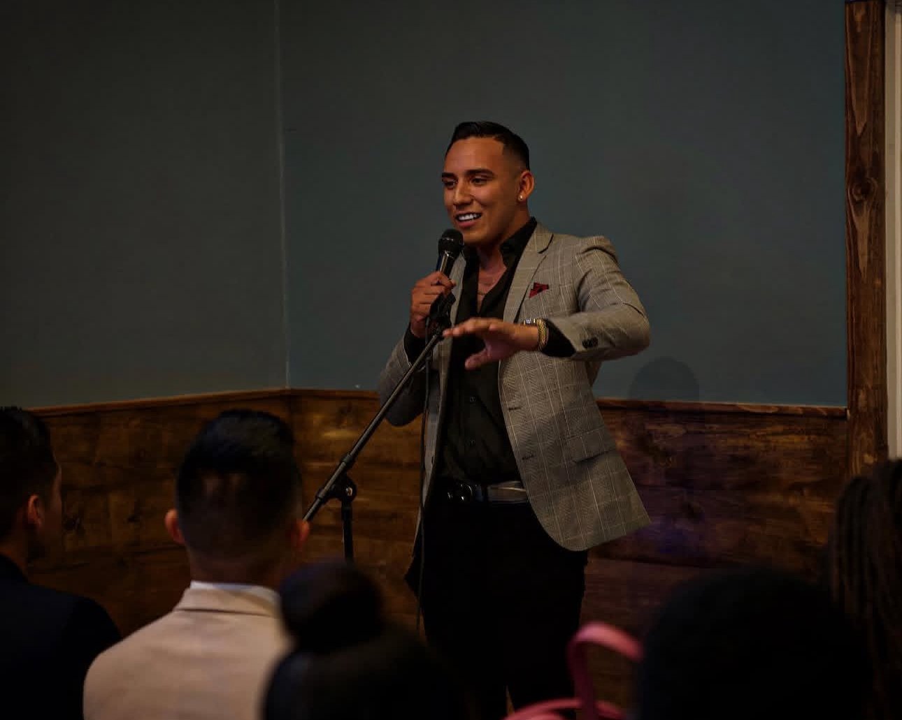 A man in a gray checked blazer and black shirt speaking into a microphone at a speaking event, with several people in the audience in front of him.