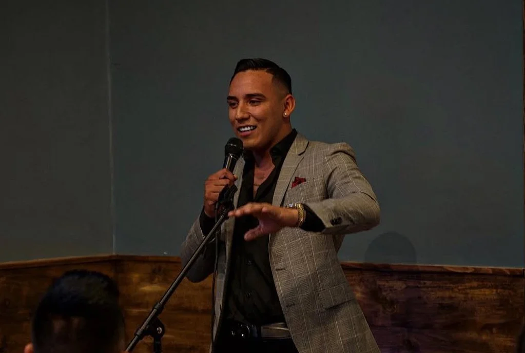 Man in gray plaid blazer speaking into a microphone in a dimly lit room