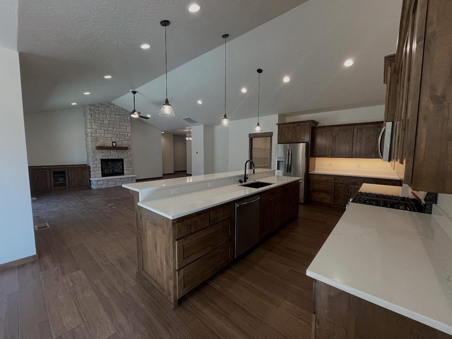 Kitchen with wooden cabinets, white countertops, stainless steel appliances, pendant lights, and a bar area, leading to a living room with a white brick fireplace and wooden flooring.