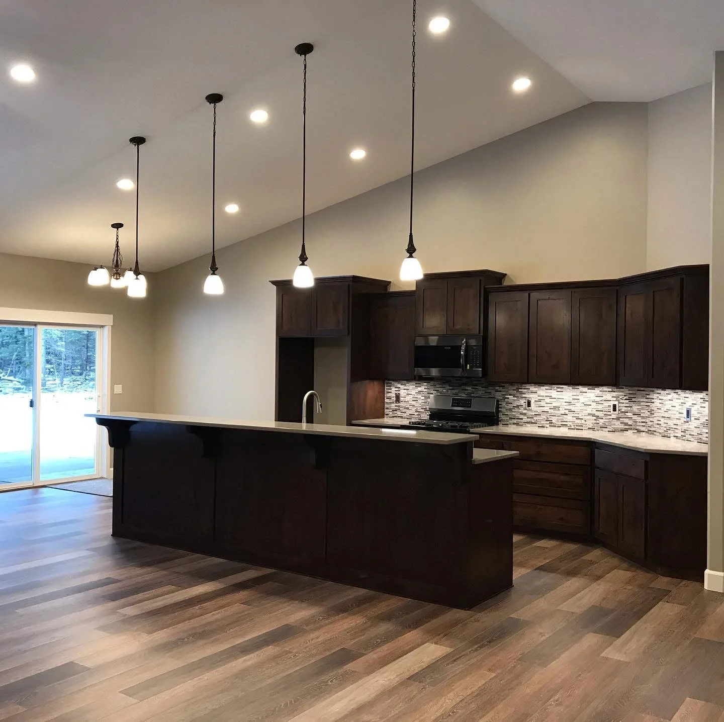Modern kitchen with dark wood cabinets, a long kitchen island, pendant lights over the island, a mosaic tile backsplash, and a sliding glass door leading outside.