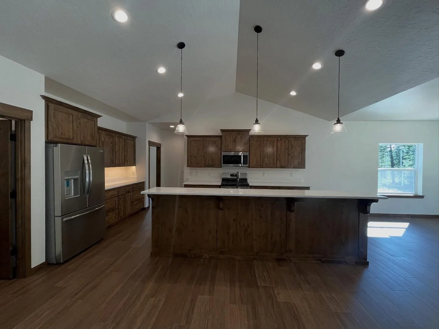 Kitchen with wooden cabinets, stainless steel refrigerator, island with white countertop, and pendant lighting, hardwood floors, and a window letting in natural light.