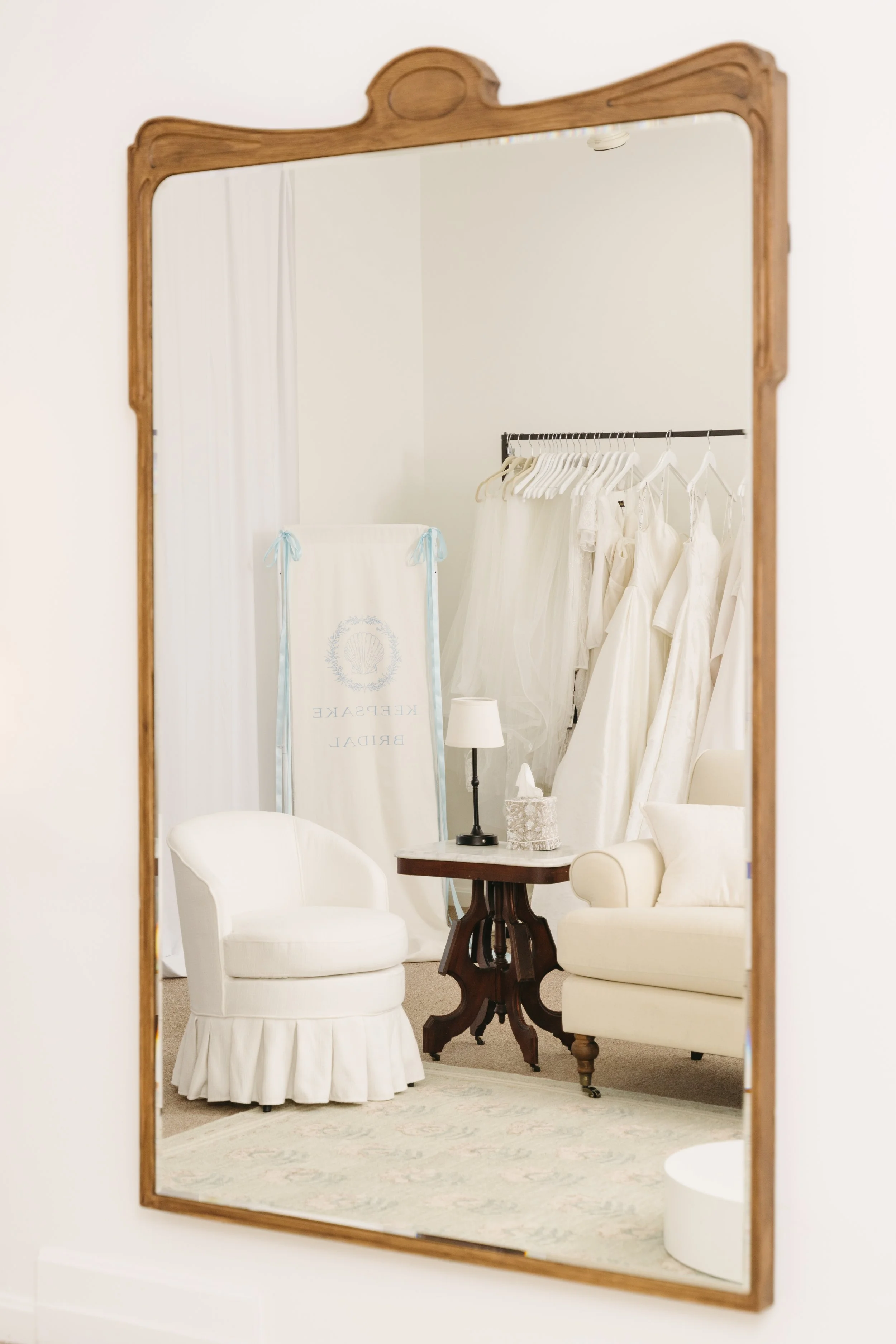 A large mirror reflects a cozy sitting area with white furniture and wedding dresses hanging on a rack, suggesting a bridal shop or fitting room.
