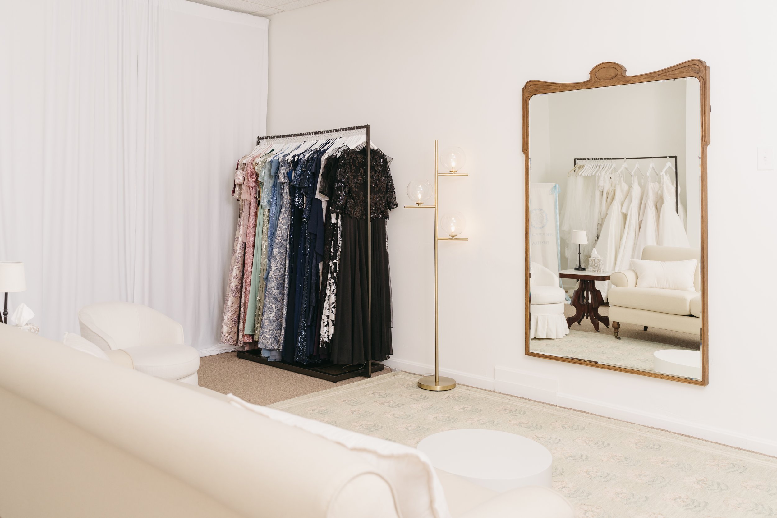 A boutique fitting room with a clothing rack holding dresses of various colors and patterns, a large mirror with a wooden frame, a white sofa, a golden floor lamp, and a cream-colored rug.