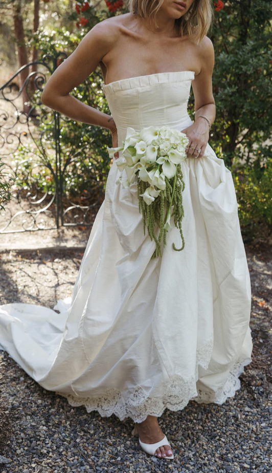 Woman wearing a strapless white wedding dress holding a bouquet of white flowers outdoors.