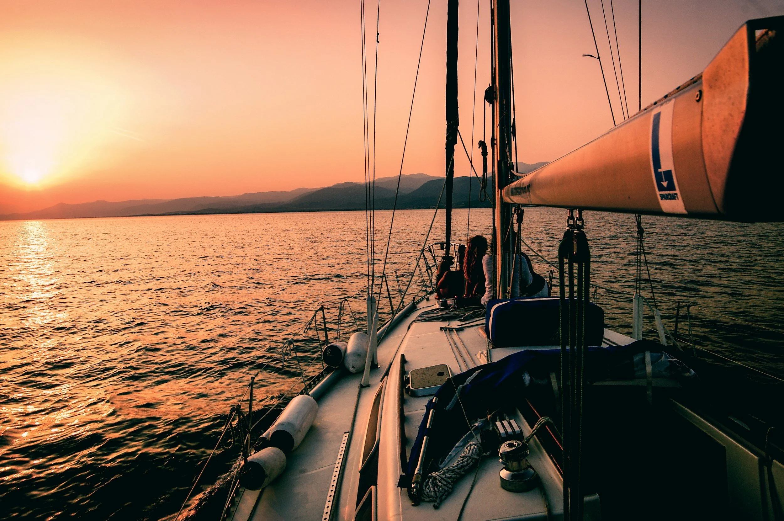 View from a sailboat during sunset, with the boat's deck and rigging visible, surrounded by calm water, and mountains on the horizon.