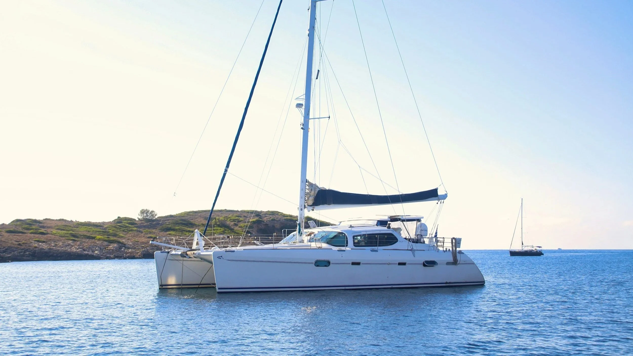 A white sailing yacht with a black mast sail anchored in calm blue water near a hilly coastline, with another sailboat in the distance under a clear sky.