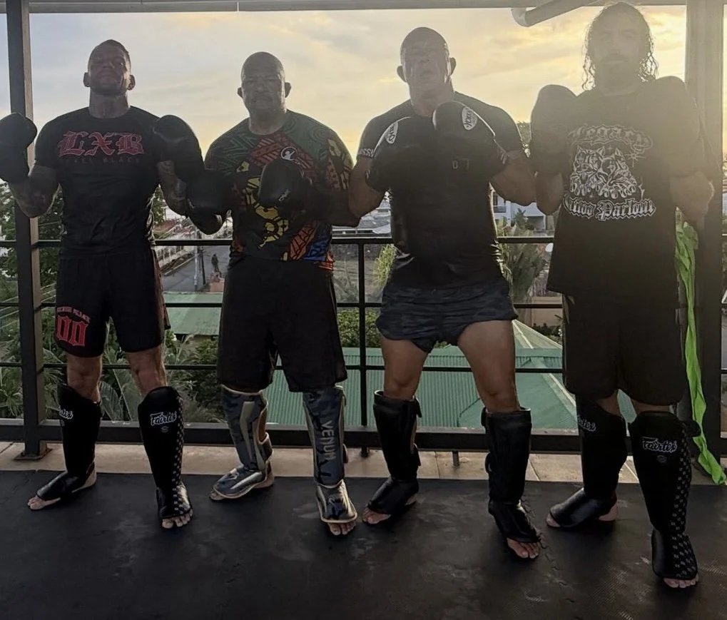Four men in boxing gear and shorts posing on a balcony with a city view at sunset.