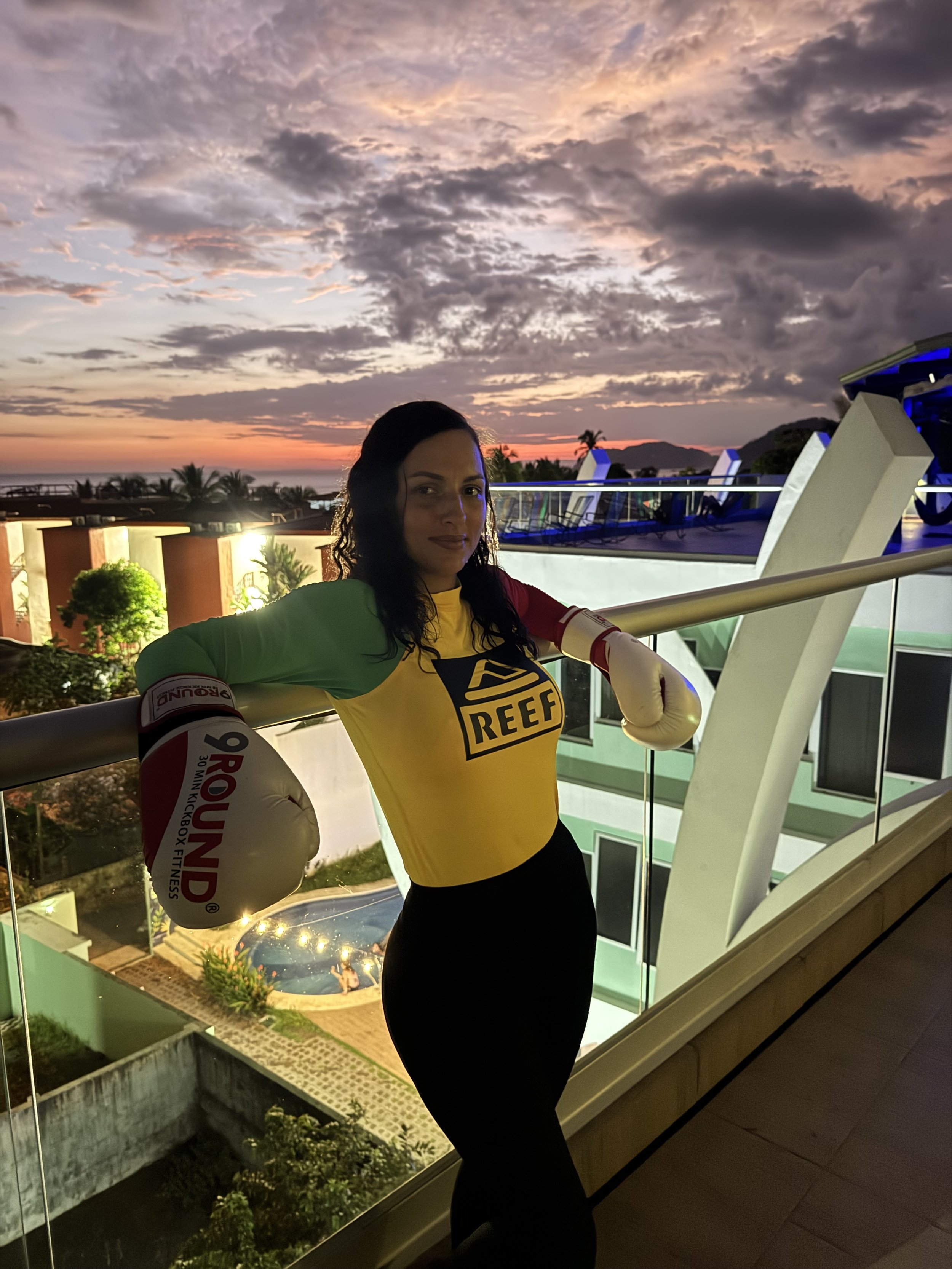 A woman with wet hair wearing a yellow Reef shirt and boxing gloves is posing on a balcony during sunset, with a scenic view of palm trees, buildings, and mountains in the background.