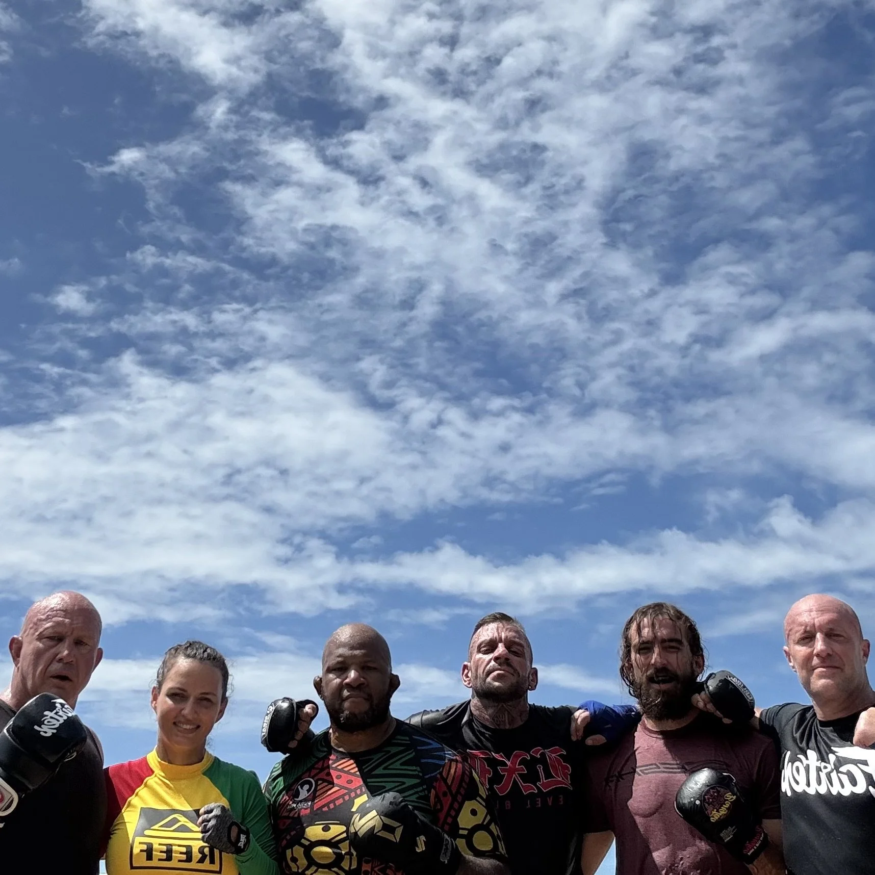 A group of diverse people wearing boxing gloves poses outdoors under a blue sky with scattered clouds, suggesting they are participating in a fitness or martial arts event.