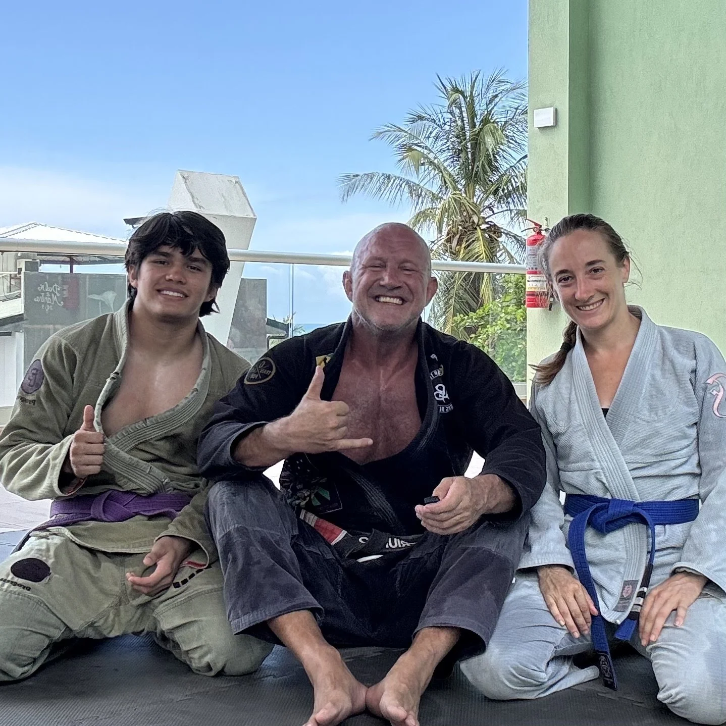 Three people in martial arts uniforms sitting and kneeling on a mat outdoors, smiling at the camera, with a palm tree and blue sky in the background.