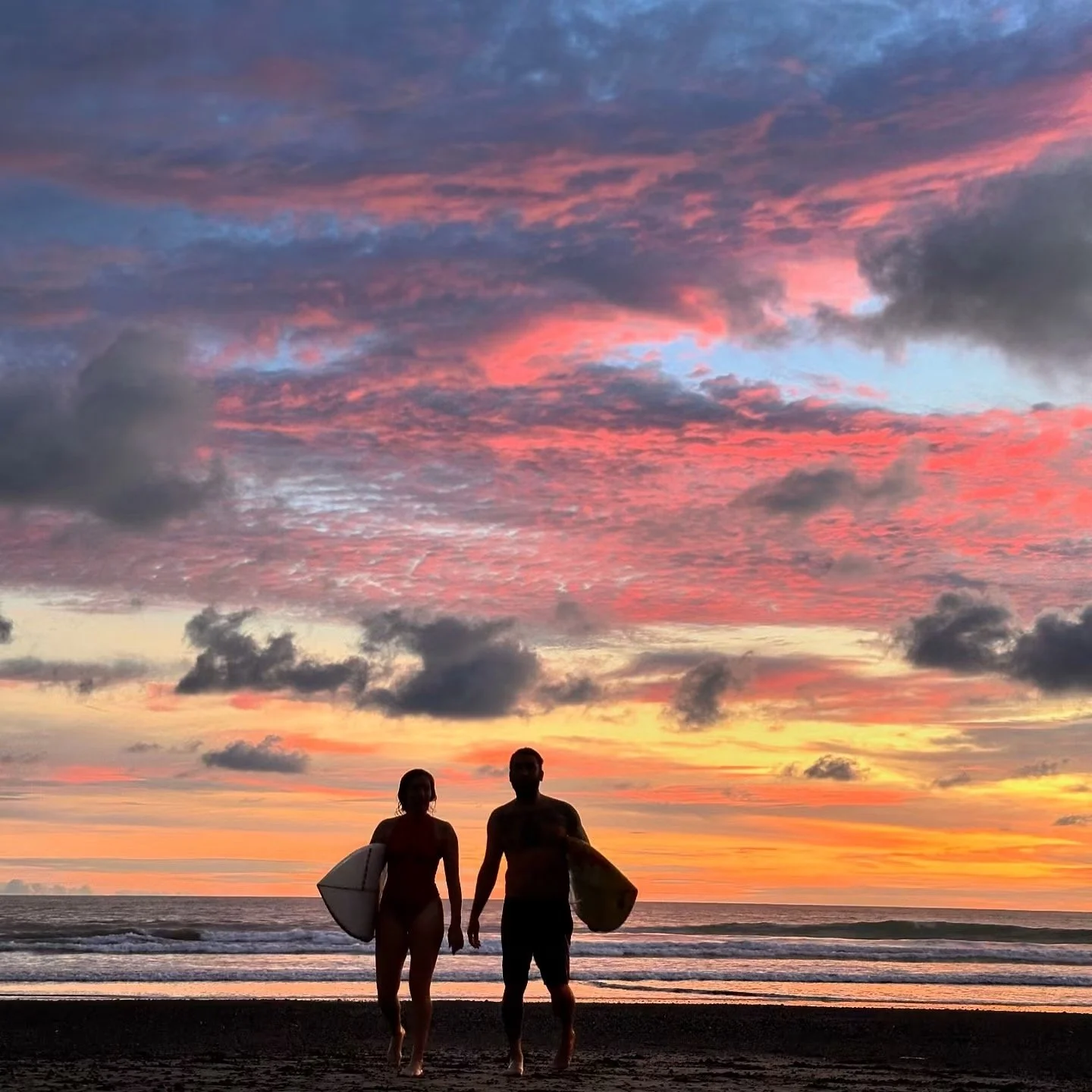 A silhouette of a man and woman walking on the beach during sunset, carrying surfboards, with colorful clouds in the sky.