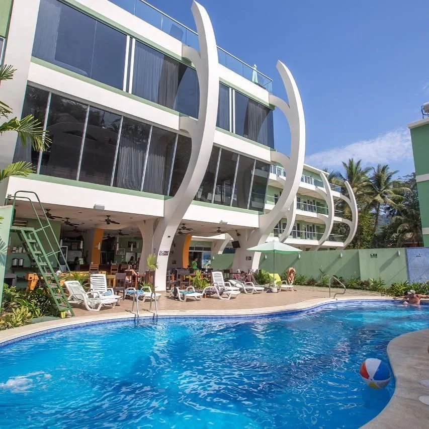 View of a tropical hotel with a swimming pool in the foreground, modern building with curved architecture, lounge chairs, and palm trees under a blue sky.
