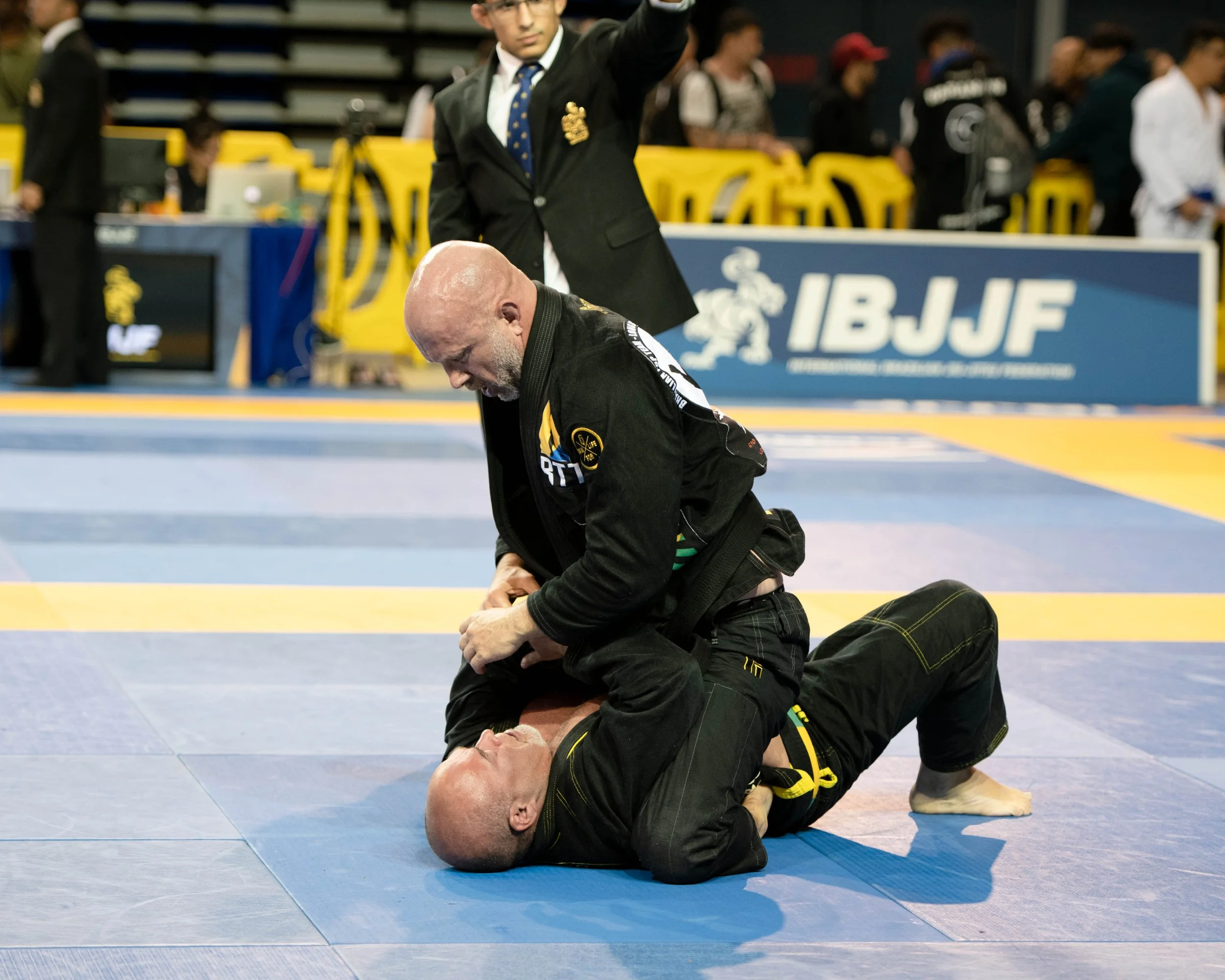 Two martial artists practicing Brazilian Jiu-Jitsu on a blue mat, with a referee in a black suit and blue tie observing in the background.