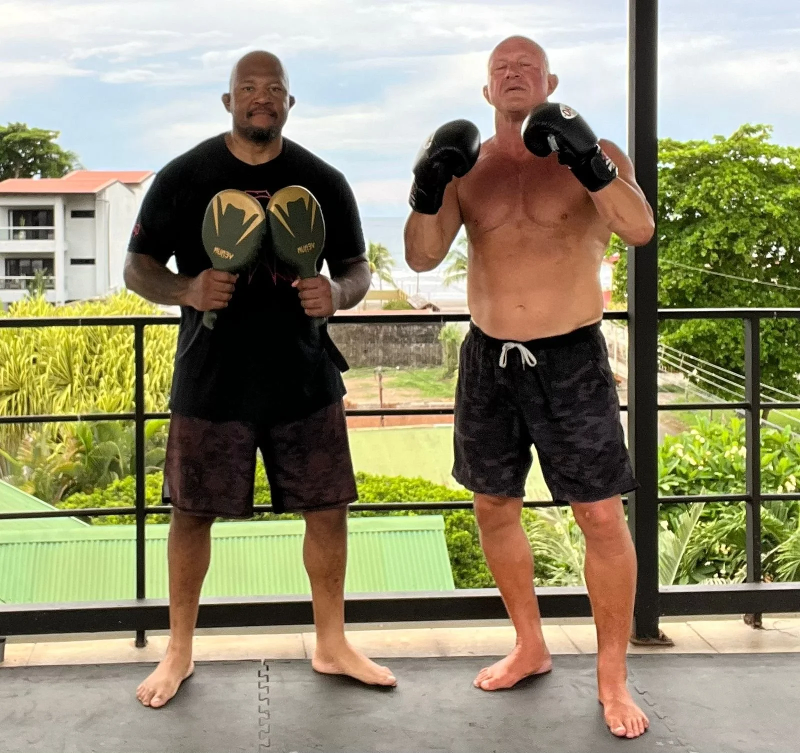 Two muscular men standing barefoot on a mat on a balcony, wearing boxing gloves and shorts. One man is shirtless with black shorts, holding up fists, the other man is wearing a black t-shirt and shorts, holding his boxing gloves near his chest. The background shows a blue sky, green trees, and some residential buildings.