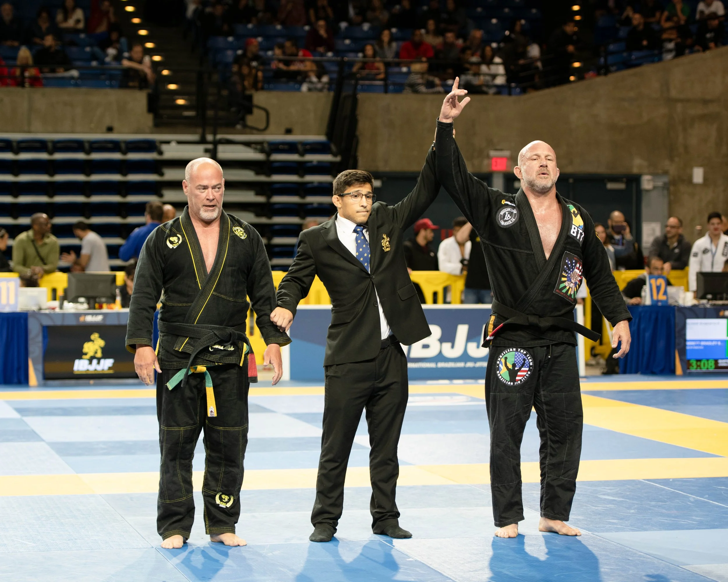 Two male Brazilian Jiu-Jitsu competitors in black gis walking off the mat, one with a yellow belt, accompanied by a referee in a suit raising the hand of the competitor with the longer beard, indicating he is the winner. The audience is seated in the background at an indoor sports arena.