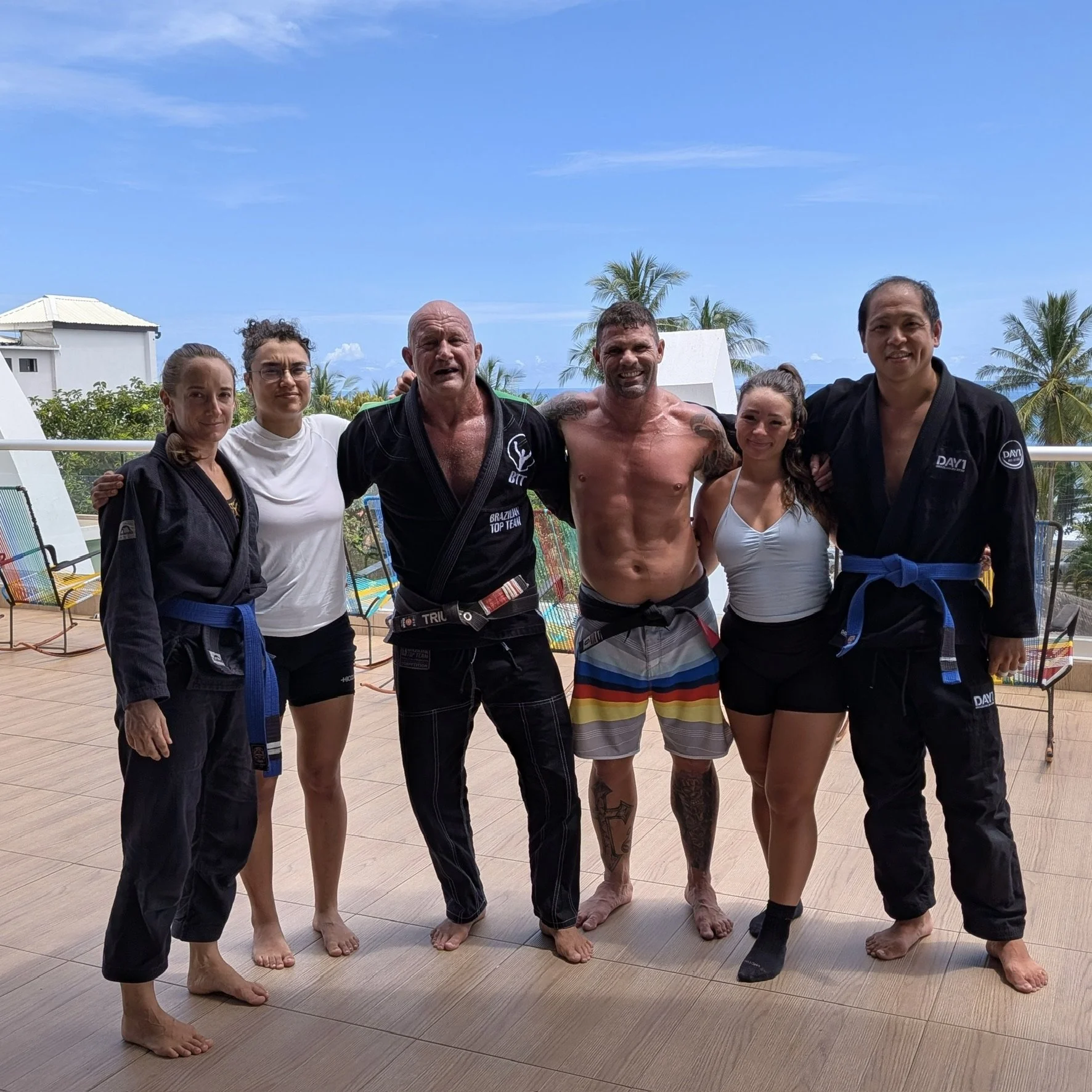 Group of six people, four women and two men, in martial arts uniforms and swimwear, standing on a deck with beach chairs and palm trees, smiling after martial arts training, under a blue sky.