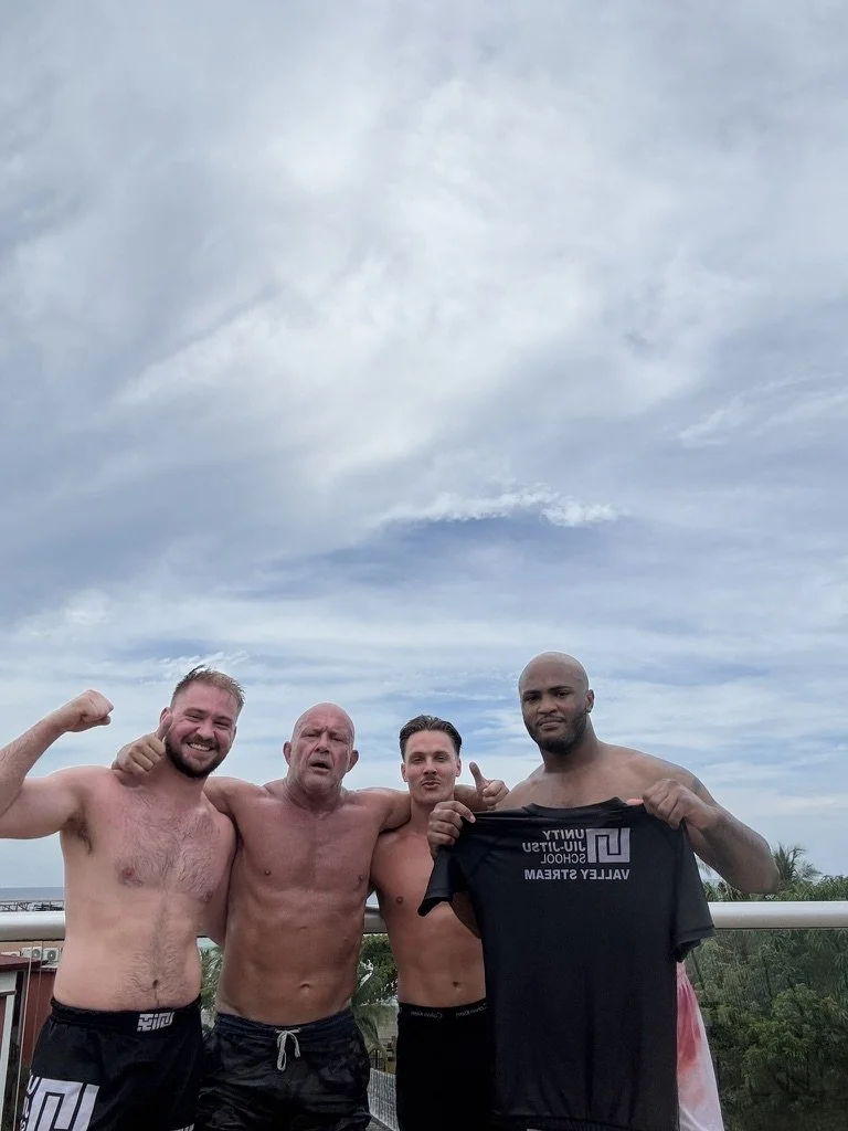 Four men shirtless on a balcony with a cloudy sky in the background, one holding a black T-shirt with white text.
