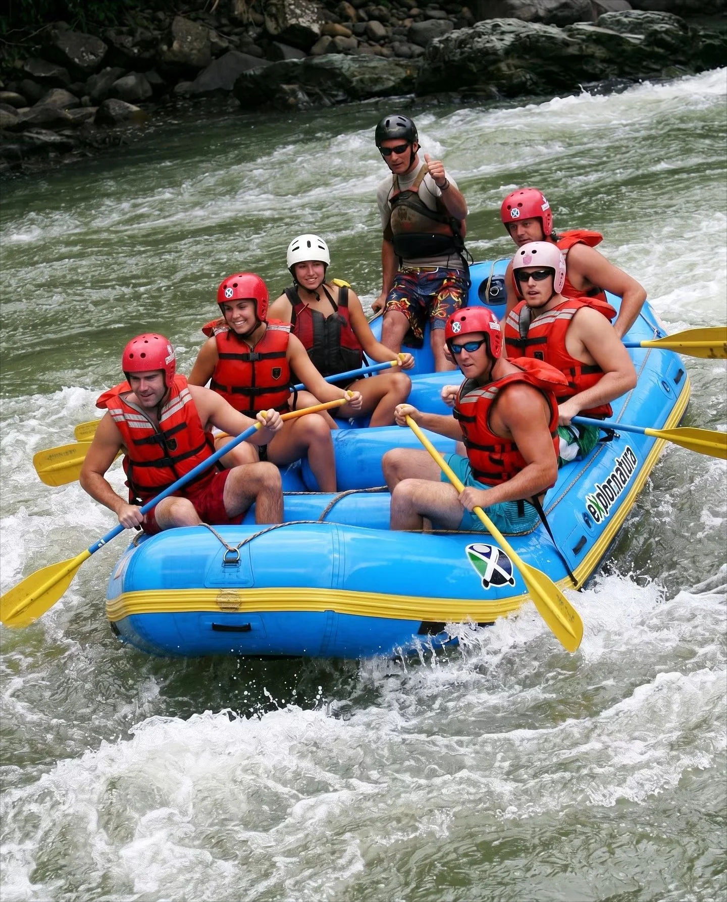 Group of seven people on a blue inflatable raft white water rafting. All are wearing life jackets, helmets, and holding paddles. They are paddling through a river with rocks and rapids, with a rocky shoreline in the background.