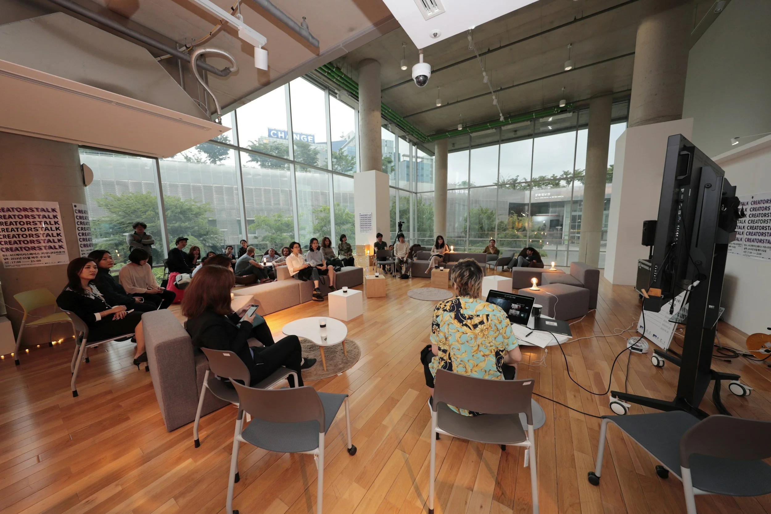 A group of people attending a presentation or discussion forum in a spacious, modern, glass-walled room with wooden floors. The presenter is sitting at a desk with a laptop, and the audience is seated on sofas and chairs, listening attentively.