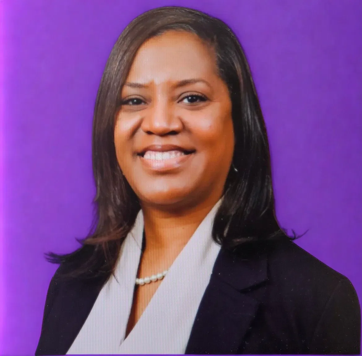 A professional woman with shoulder-length black hair, wearing a black blazer over a white blouse with a pearl necklace, smiling against a purple background.