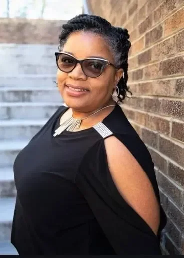 A woman with dark hair styled in twists, wearing sunglasses, a black top with cut-out shoulders, and a white and gold necklace, standing against a brick wall outdoors.