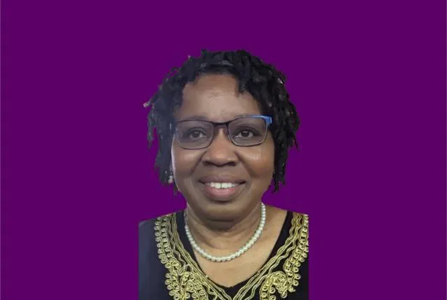 Portrait of a woman with glasses, short curly hair, wearing a black and gold embroidered top and pearl necklace, against a purple background.