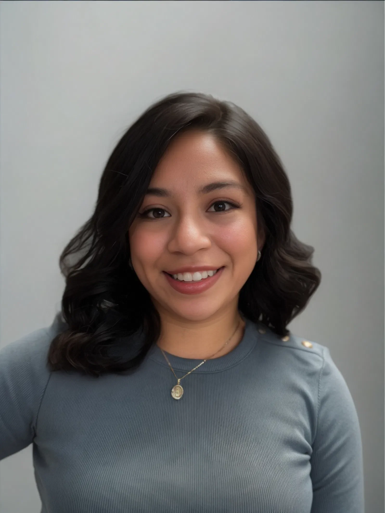 A young woman with dark, wavy hair smiling at the camera, wearing a gray top with button details on the shoulder and a gold necklace with a pendant.