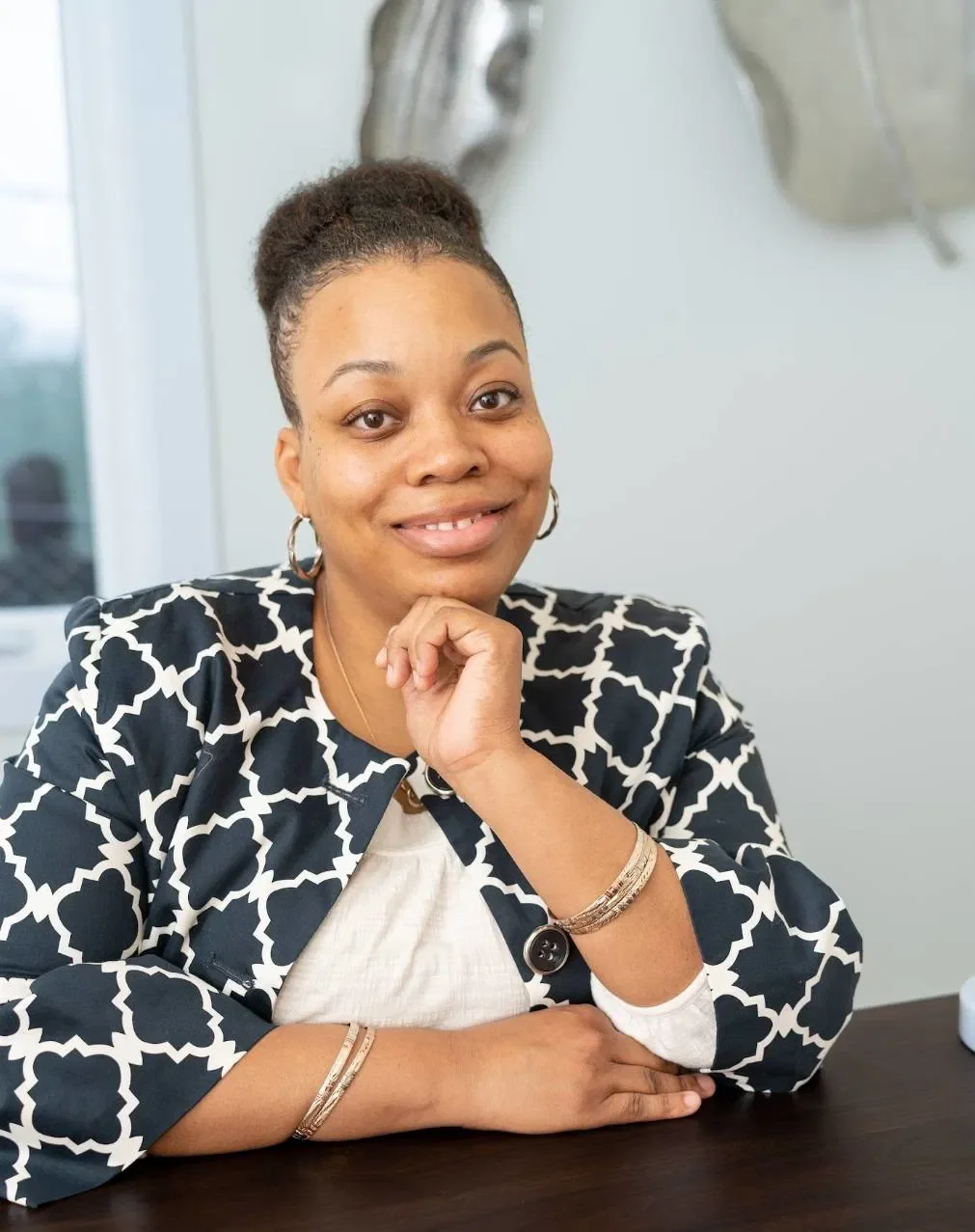 A woman with dark hair in a bun, wearing hoop earrings, a patterned blazer, and jewelry, sitting at a desk with her chin resting on her hand, smiling at the camera.