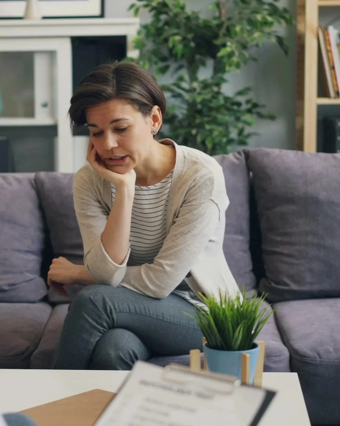 A woman sitting on a grey couch in a counseling session, looking worried or stressed, resting her head on her hand, with a potted plant on a table in front of her and bookshelves in the background.