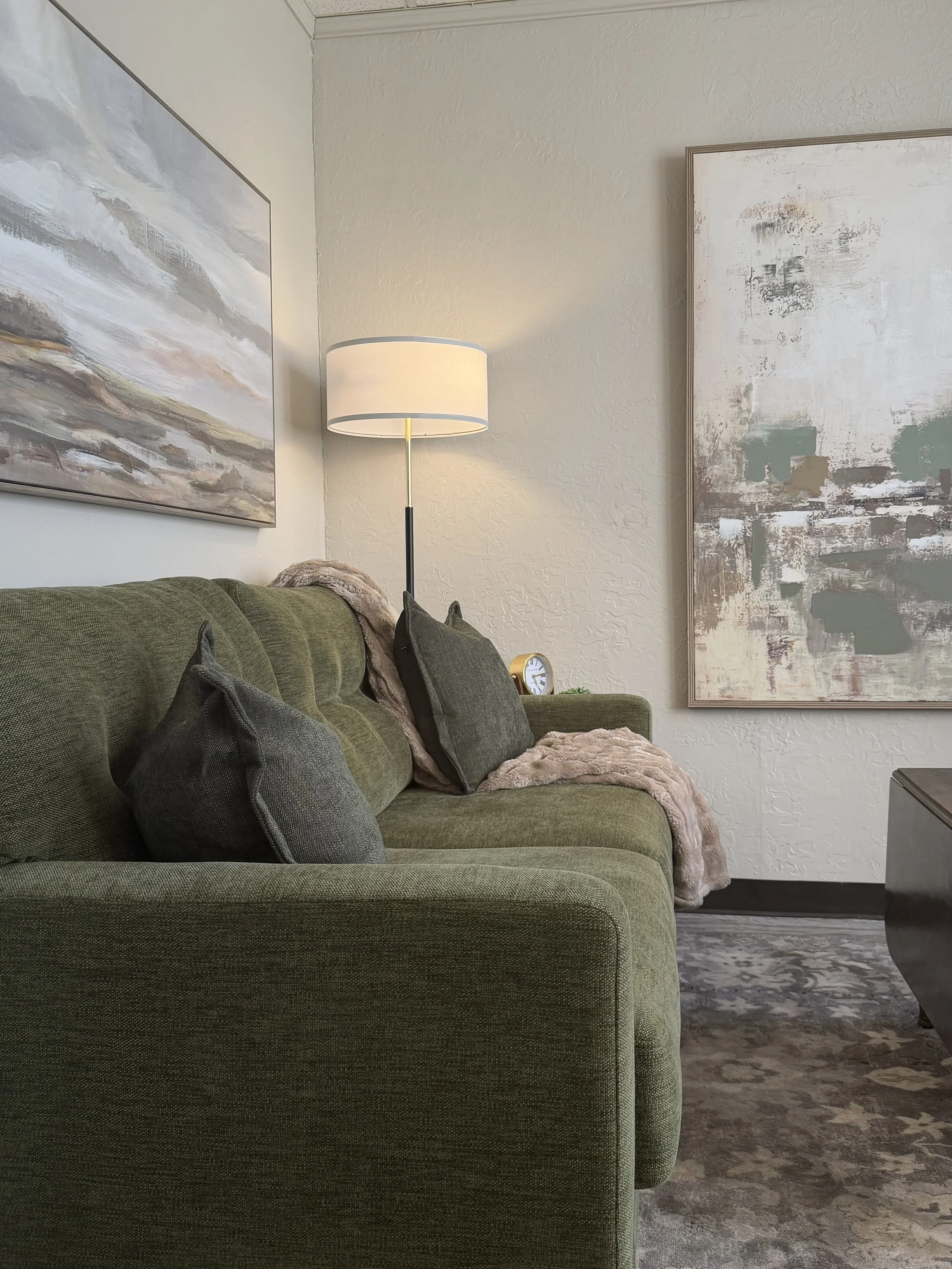 Living room corner with a green upholstered sofa, gray pillows, a pile of beige blanket, a white lampshade on a black and gold floor lamp, abstract paintings on cream-colored walls, and a patterned gray rug.