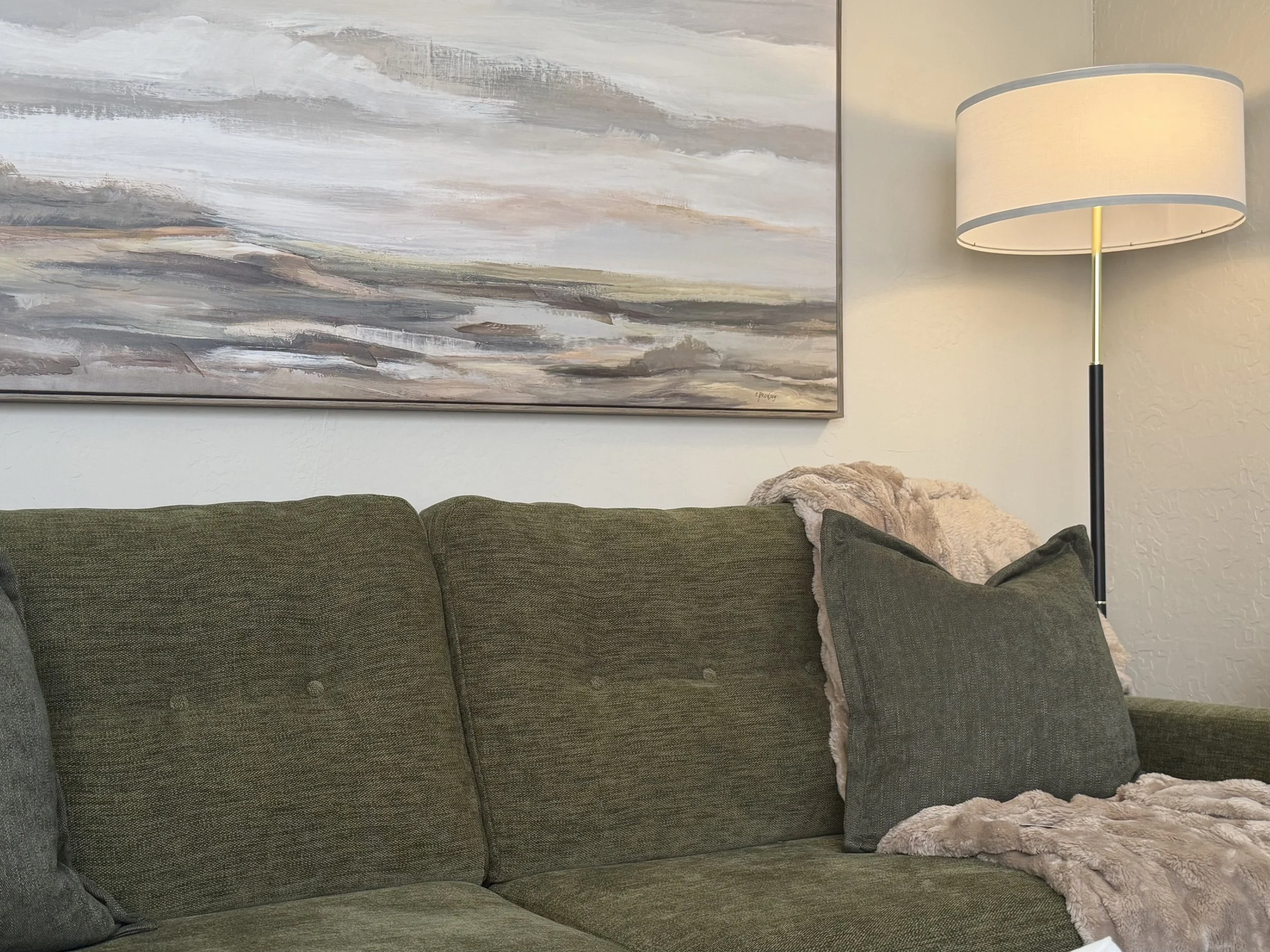 A living room with a green couch, a gray pillow, a beige throw blanket, a large abstract painting, and a tall floor lamp with a beige shade.