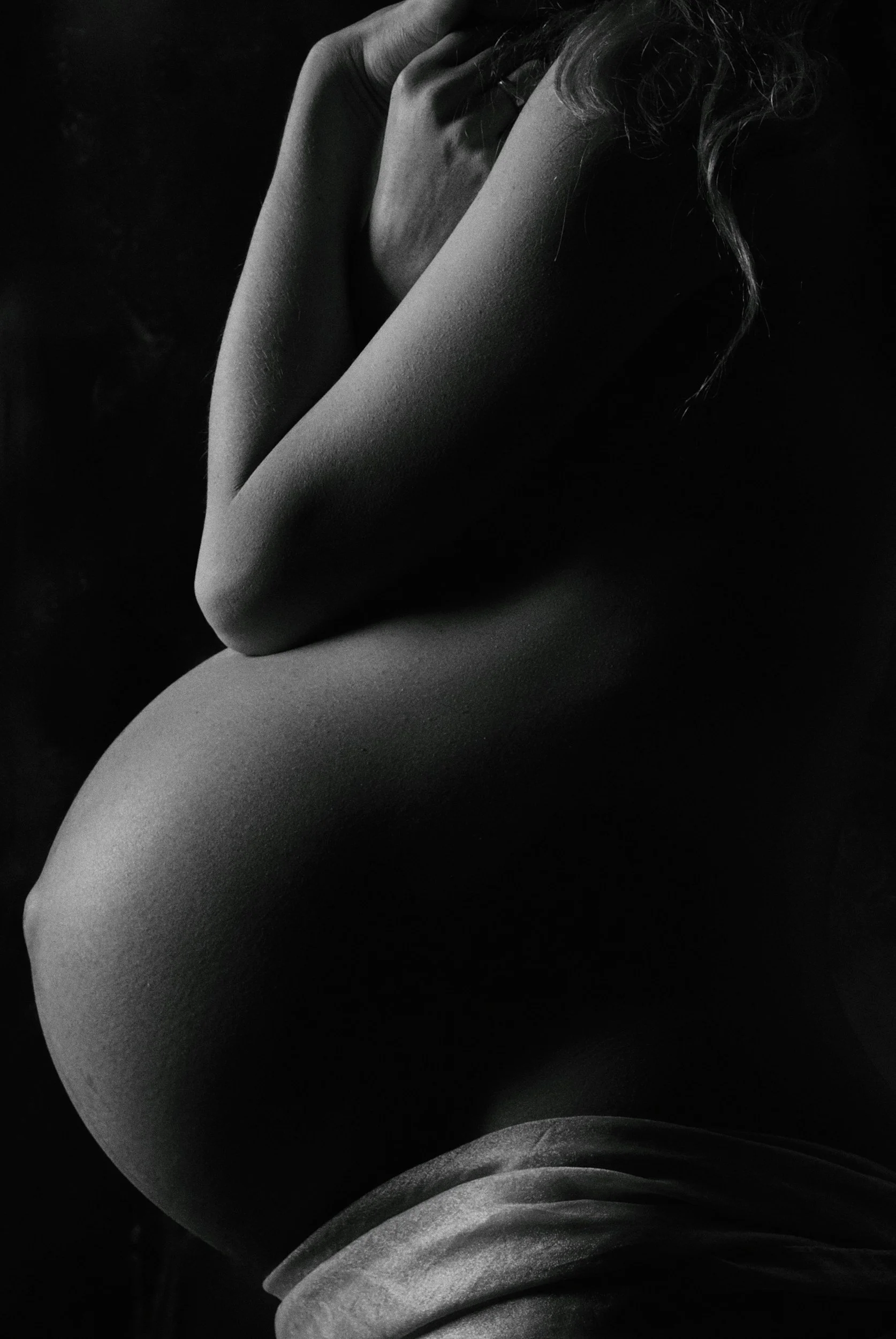 A black and white photograph of a nude woman with her body partially curled and her hand resting near her face, with curly hair and soft lighting.