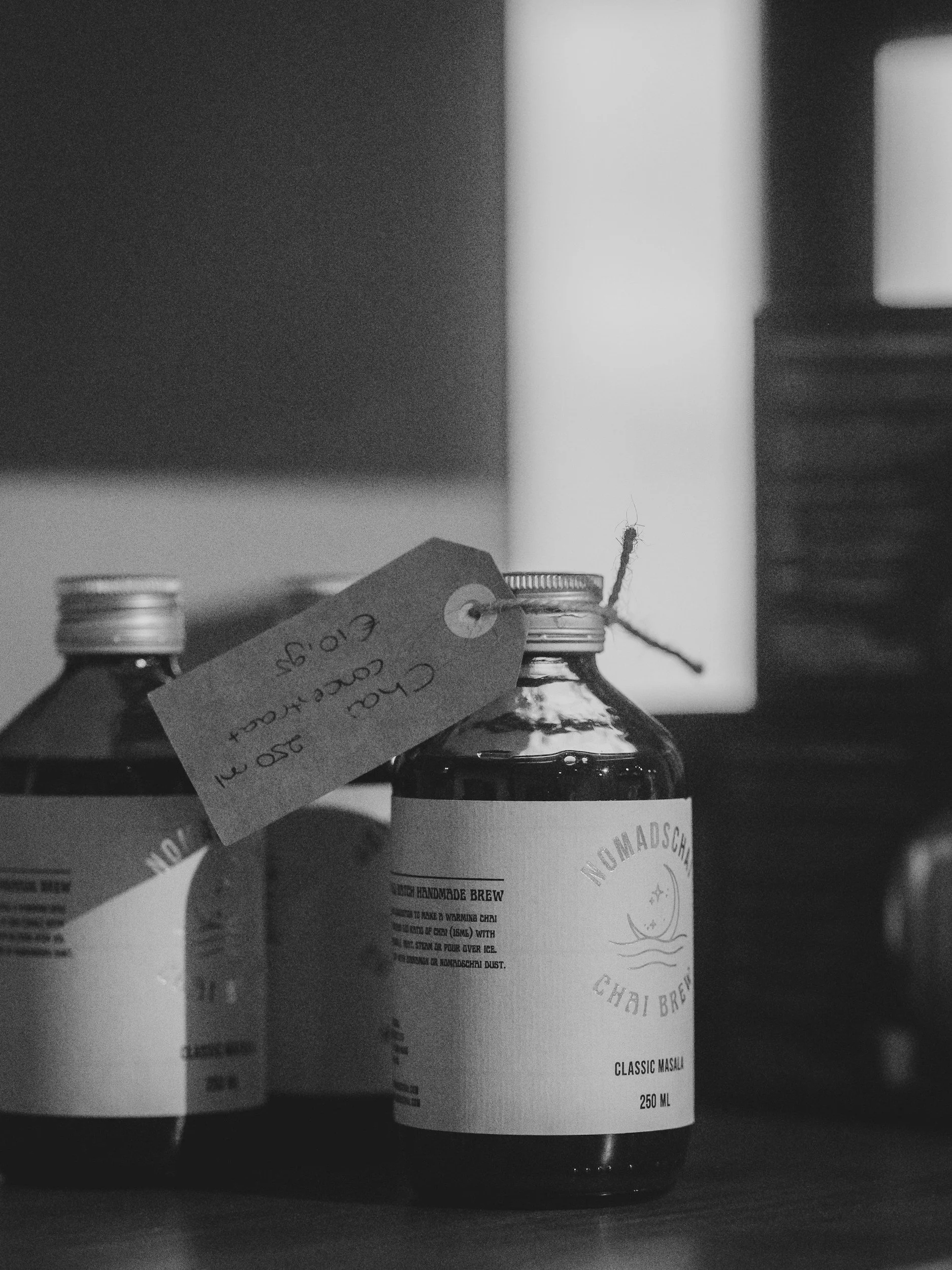 Close-up of a bottle with a label that reads 'No Madschi' and 'Classic Masaju,' with a tag tied to the neck of the bottle. The background is blurred, and the image is in black and white.