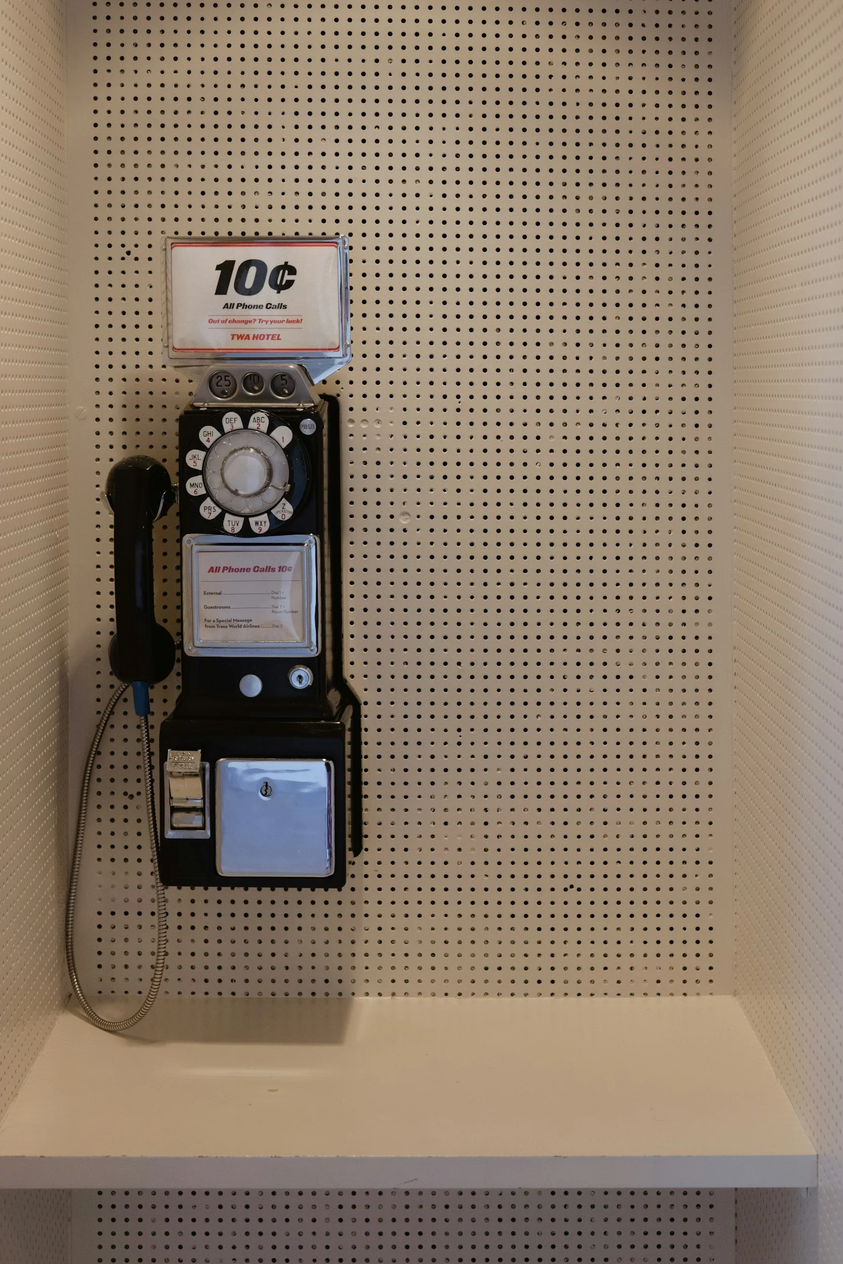 Vintage payphone with a black handset, coin slot, and keypad, mounted on a perforated beige wall.