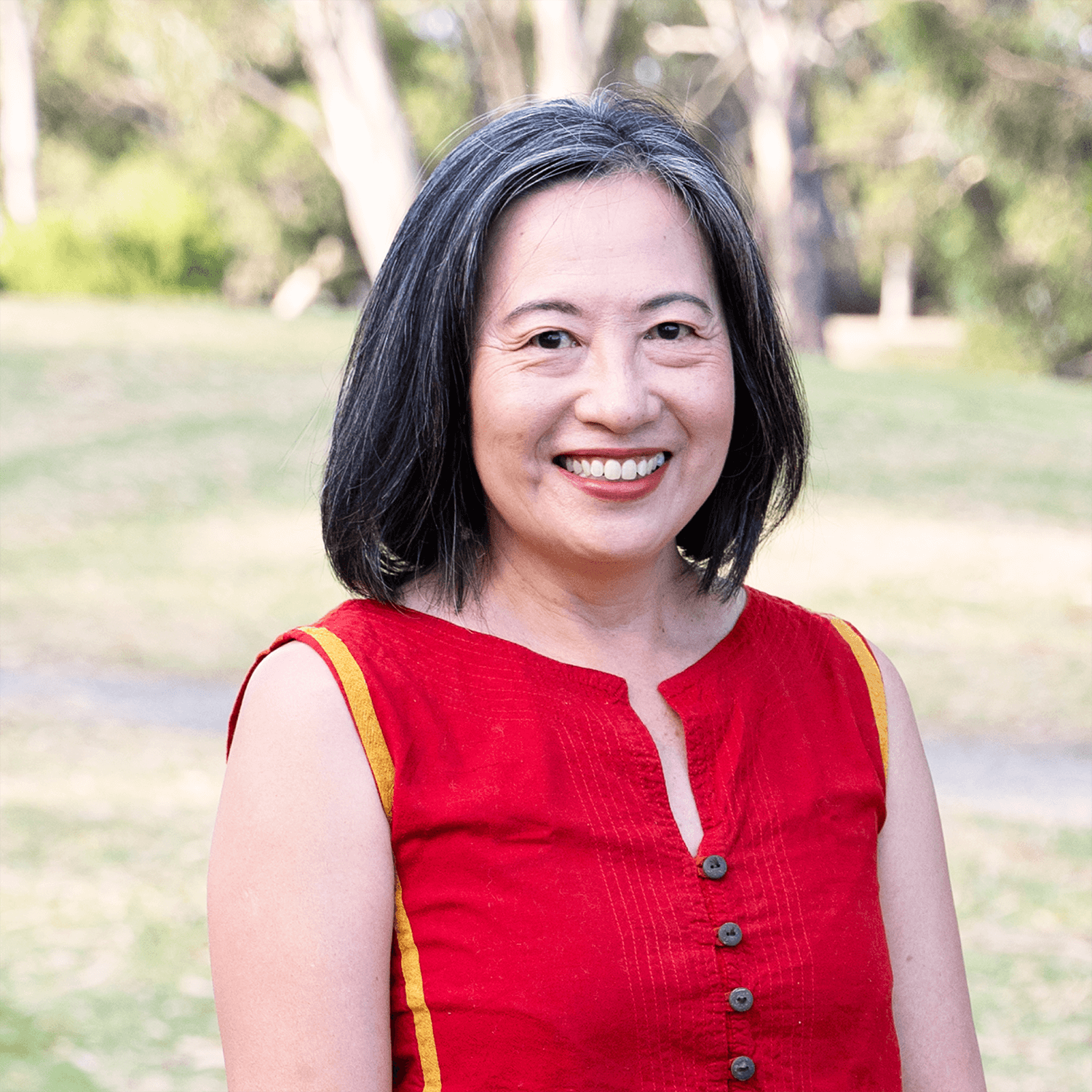 A woman with shoulder-length black hair smiling outdoors. She is wearing a sleeveless red top with yellow accents and buttons down the front. The background is a park with green grass and trees.