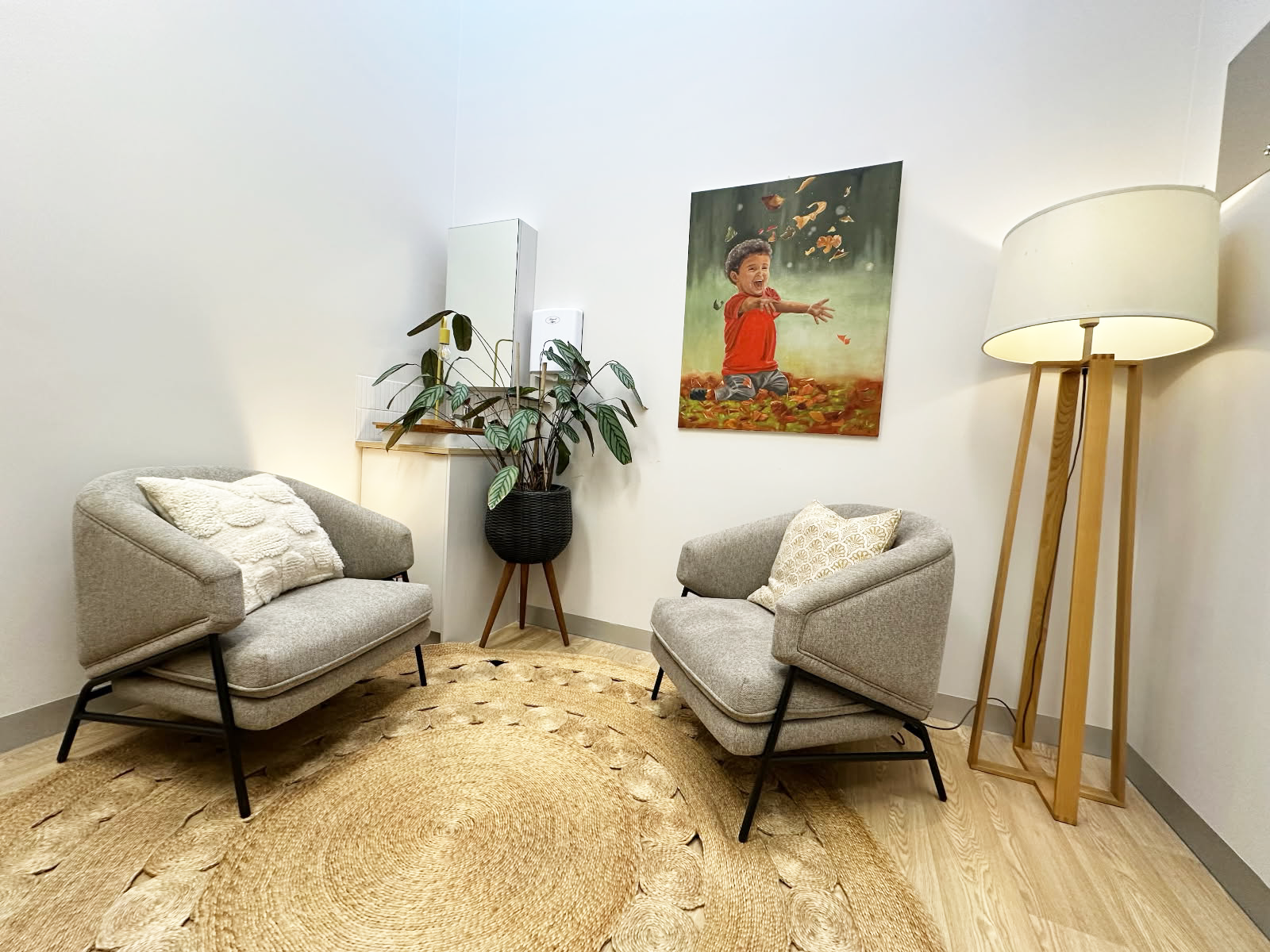 A modern living room with two light gray armchairs with pillows, a large potted plant, a floor lamp, a framed picture of a smiling child playing outdoors, a woven rug, and a small white cabinet.