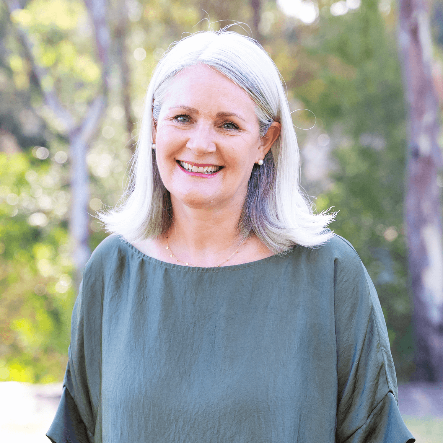 A smiling woman with silver hair and pearl earrings outdoors, wearing a green top, with a blurred background of trees and greenery.