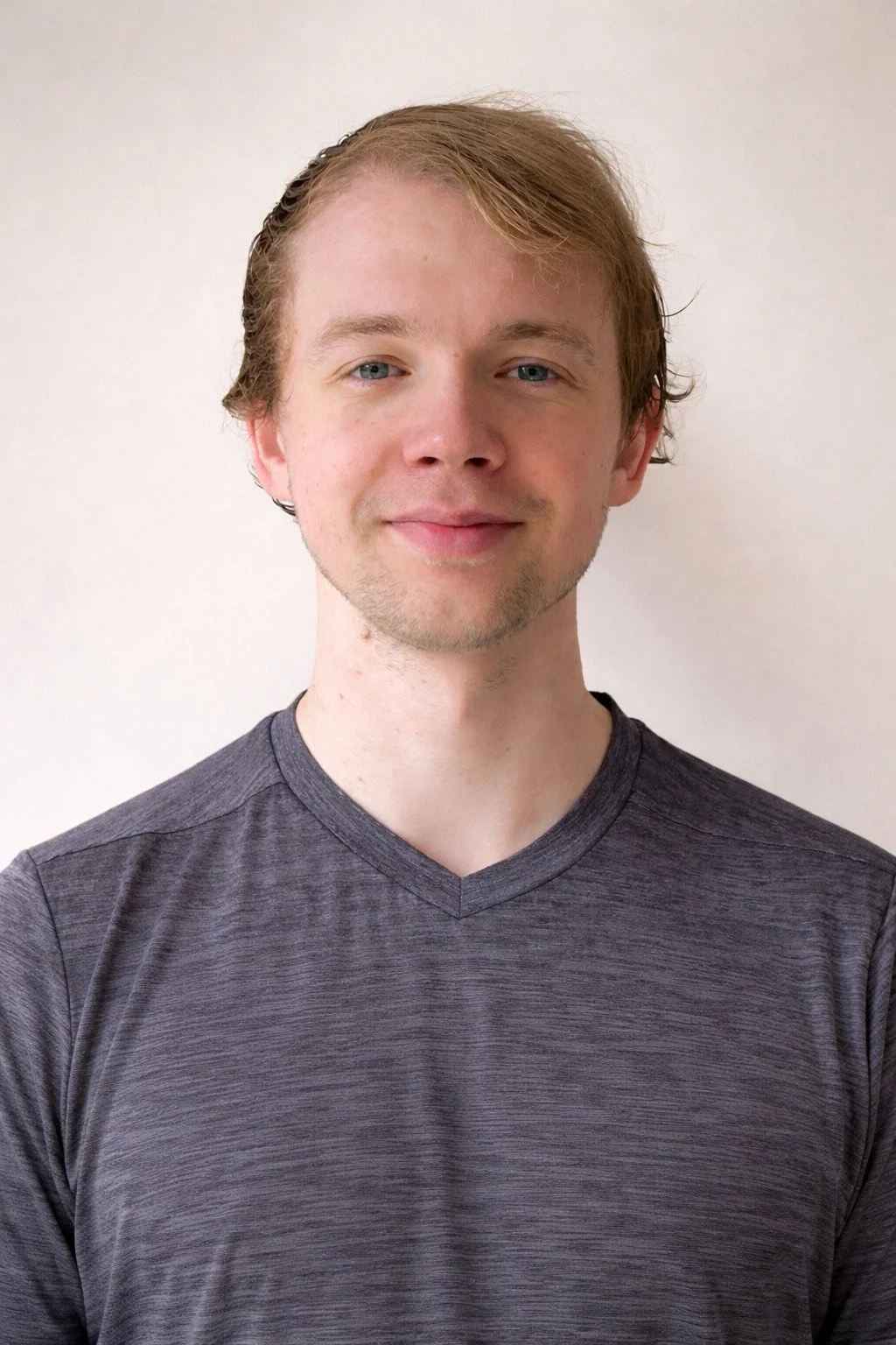 A young man with light skin, light brown hair, and blue eyes, smiling and wearing a dark gray V-neck shirt, standing against a plain white background.