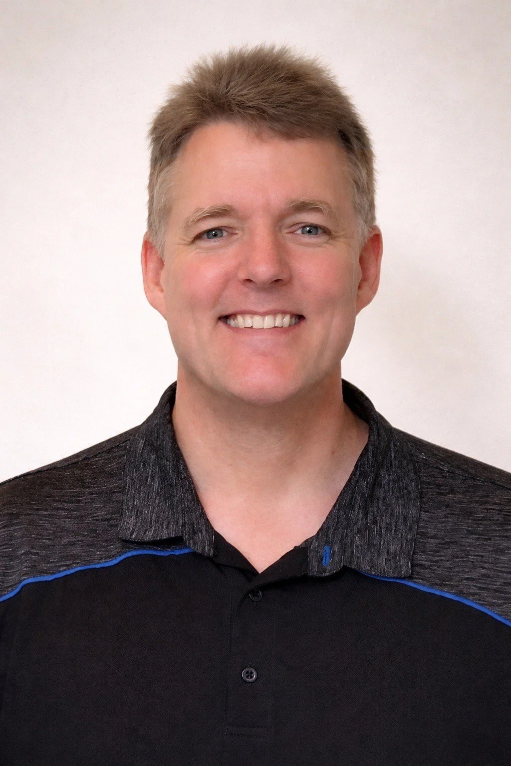 A smiling man with short light brown hair, wearing a black and gray collared shirt with blue accents, standing against a plain white background.